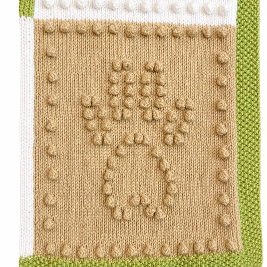 Close-up texture detail of the Dog Life baby blanket knitting pattern showing raised bobble stitch paw print motif with green, cream and beige knitted sections.