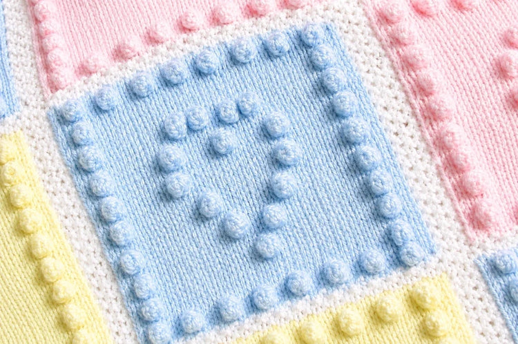 Close up of Heart Squares baby blanket knitting pattern showing pastel blue heart bobble square with soft pink and yellow knitted panels and textured white borders