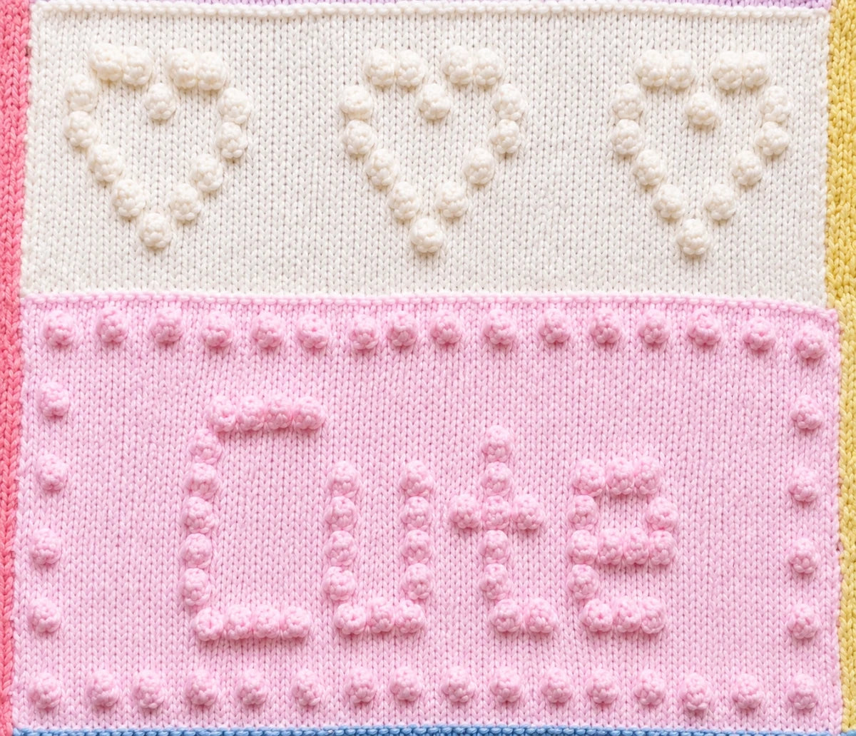 Close-up of the Sweet Dreams baby blanket showing bobble stitch hearts and the Cute word panel knitted using knit, purl and bobble stitches.