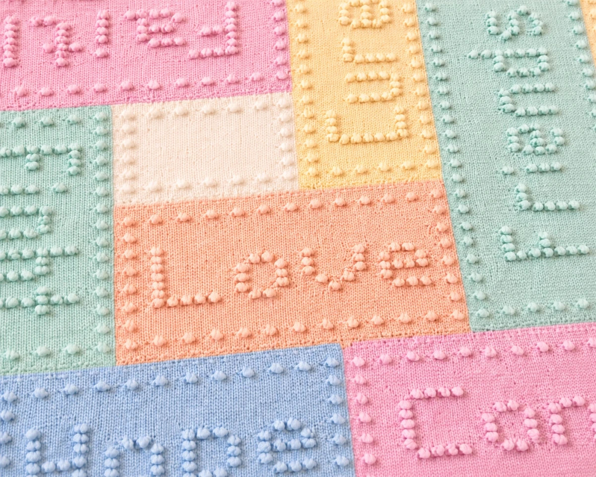 Close up of pastel cancer support lap blanket knitting pattern showing textured words and bobble stitch border detail in soft pink, blue, mint and peach panels.