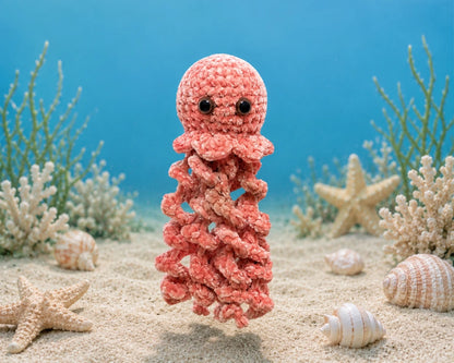 Soft pink crochet jellyfish plush toy floating above a sandy underwater scene with seashells and starfish