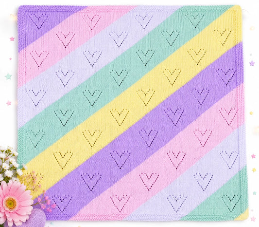 Pastel diagonal lace baby blanket knitting pattern with soft heart motifs in pink, lilac, mint and yellow stripes displayed in a bright flat lay scene with flowers and spring styling