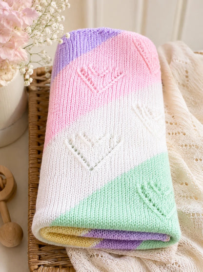 Folded pastel diagonal lace baby blanket knitting pattern with heart motifs displayed in a soft nursery scene with flowers, woven tray and cosy neutral styling