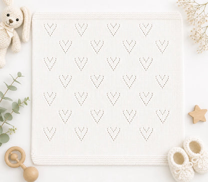 White knitted baby blanket with delicate heart lace motifs and garter stitch border displayed in a soft neutral nursery flat lay scene with bunny toy and baby accessories.