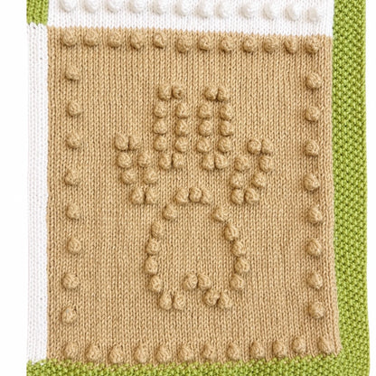 Close-up texture detail of the Dog Life baby blanket knitting pattern showing raised bobble stitch paw print motif with green, cream and beige knitted sections.
