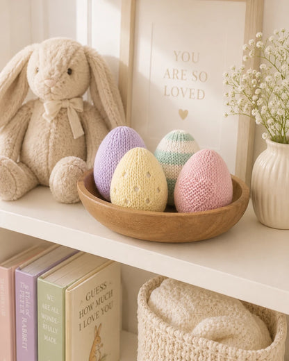 Pastel knitted Easter eggs displayed in a wooden bowl on a nursery shelf beside a soft bunny teddy and pastel nursery decor from a free Easter knitting pattern bundle by Peach Unicorn Designs