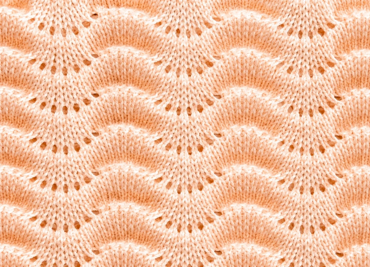 Close up texture detail of the Gentle Waves baby blanket knitting pattern in soft peach yarn showing the repeating scalloped lace wave stitch design.