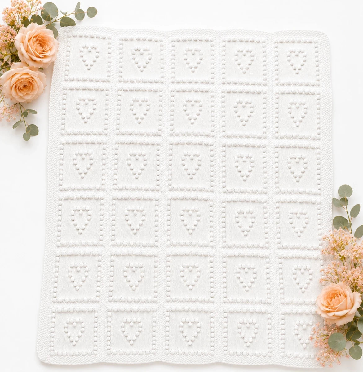 All white Heart Squares baby blanket knitting pattern with textured bobble heart squares on a soft white background with peach flowers and eucalyptus styling