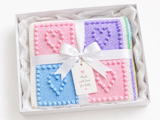 Gift wrapped Heart Squares baby blanket knitting pattern with pastel bobble heart squares, white ribbon and a gift tag reading “From my heart to yours” on a soft white background