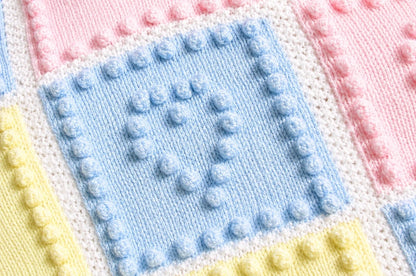 Close up of Heart Squares baby blanket knitting pattern showing pastel blue heart bobble square with soft pink and yellow knitted panels and textured white borders
