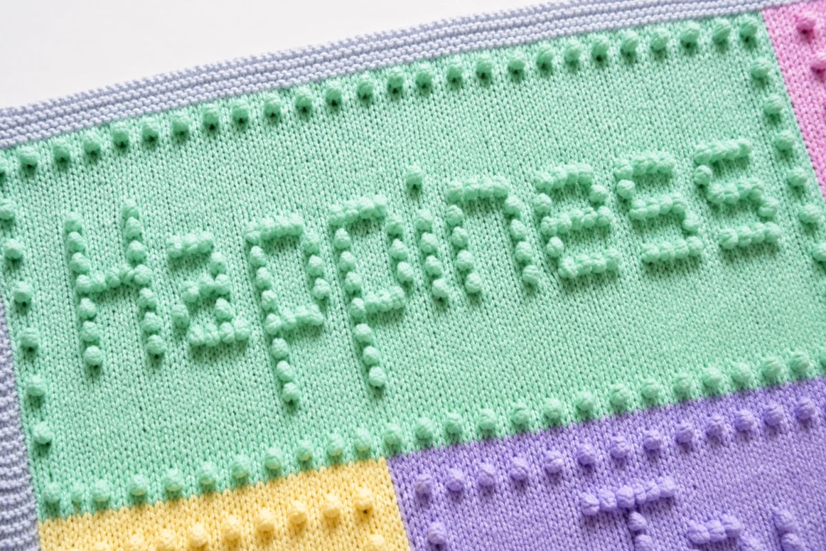Close-up of the Happiness panel on the Joy baby blanket knitting pattern showing raised bobble lettering and textured intarsia knitting