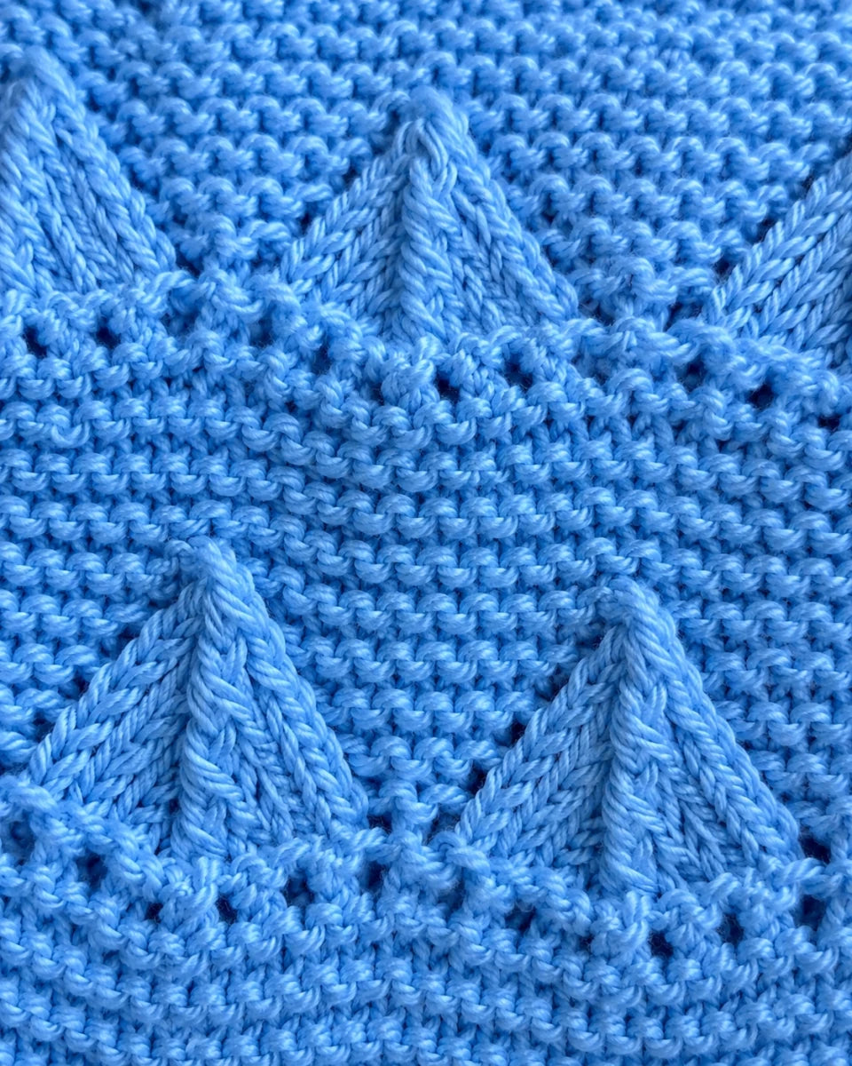 Elegant close up of the Little Boats baby blanket knitting pattern showing raised blue boat motifs, textured garter stitch rows and delicate eyelet details in soft baby blue yarn