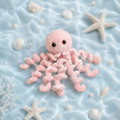 Soft pink plush jellyfish knitting pattern displayed on pale blue fabric with seashells and starfish in an underwater inspired ocean scene