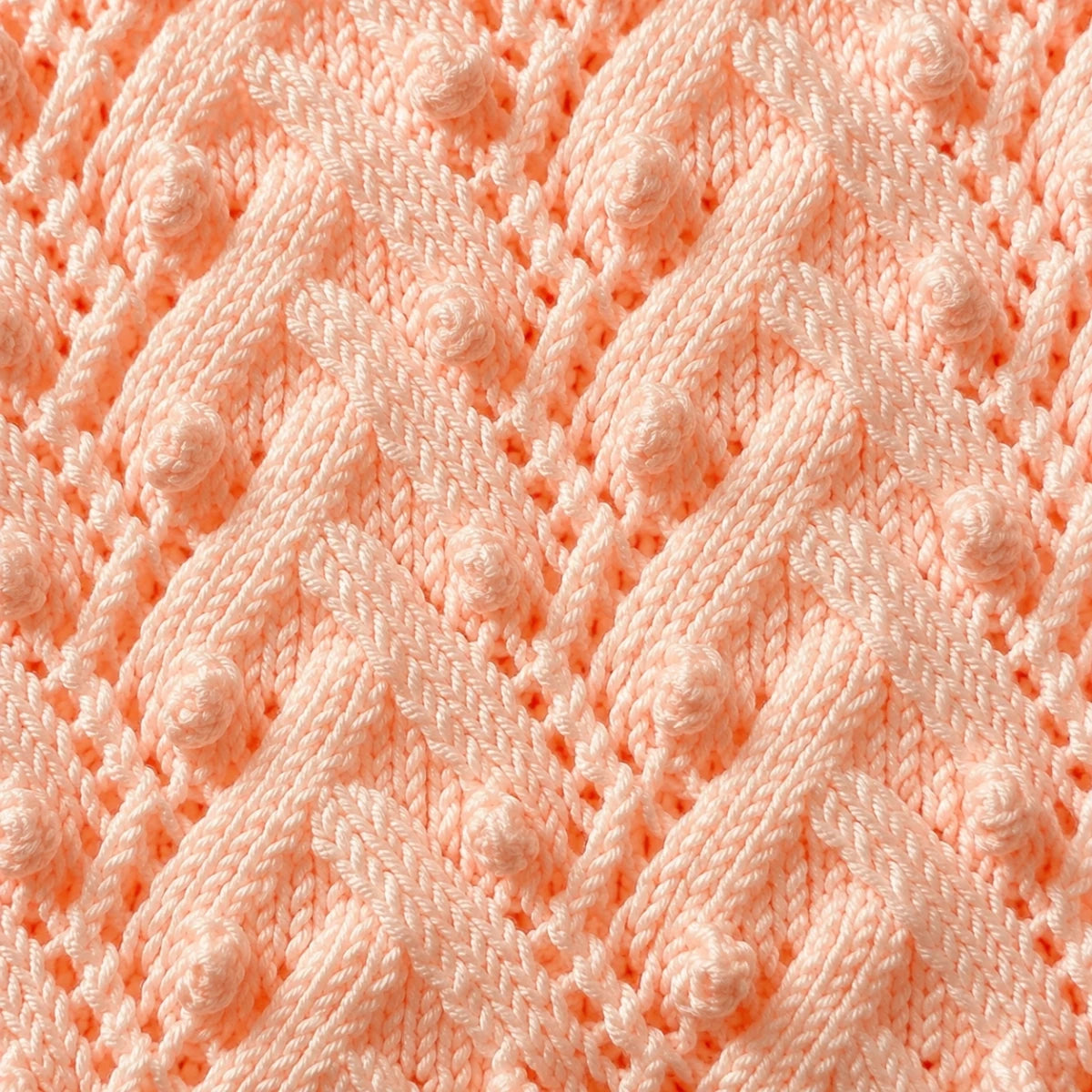 Elegant close up of peach Plaited Bobbles baby blanket knitting pattern showing textured plaited cables, lace details and raised bobble stitches in soft pastel yarn