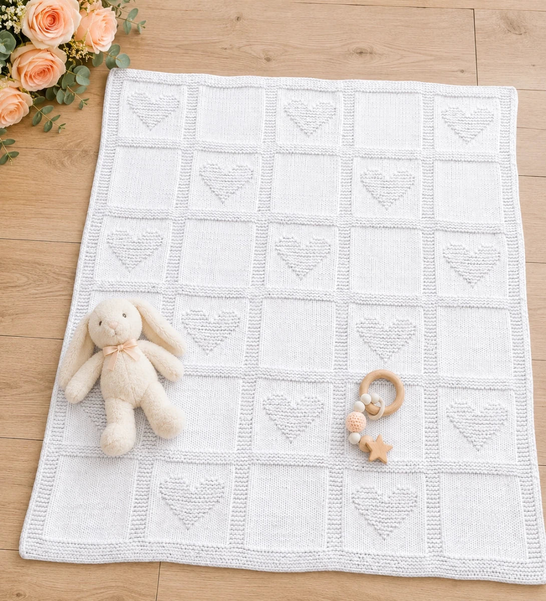 Simple Hearts Baby Blanket knitting pattern displayed on a light wooden nursery floor with peach roses, plush bunny toy and wooden baby teether, featuring alternating heart and plain knit squares.