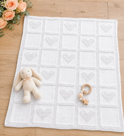 Simple Hearts Baby Blanket knitting pattern displayed on a light wooden nursery floor with peach roses, plush bunny toy and wooden baby teether, featuring alternating heart and plain knit squares.