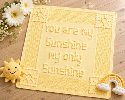 You Are My Sunshine baby blanket knitting pattern shown in soft baby yellow on a wooden floor with white flowers and sunshine-themed baby toys