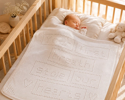 Sleeping baby covered with the Your First Breath Stole Our Hearts baby blanket knitting pattern in a wooden cot with soft morning light