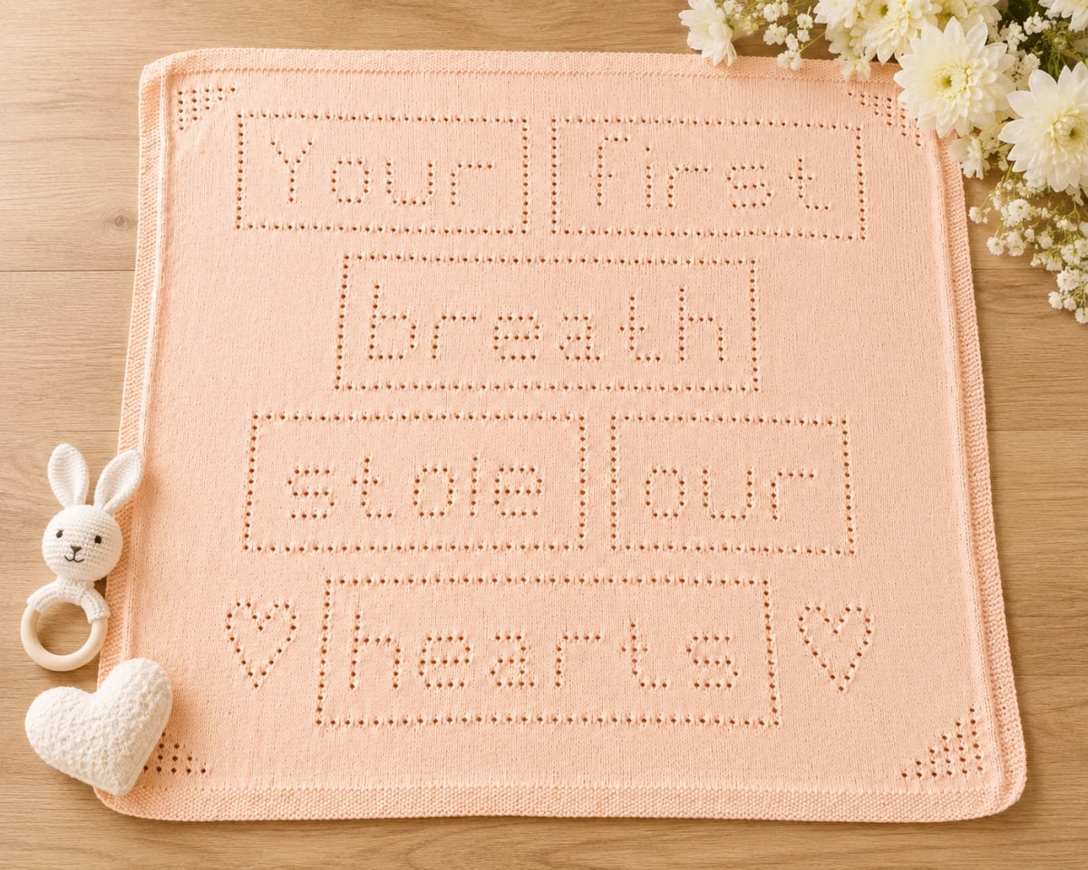 Your First Breath Stole Our Hearts baby blanket knitting pattern shown in soft peach on a wooden nursery floor with white flowers, heart toy and bunny rattle